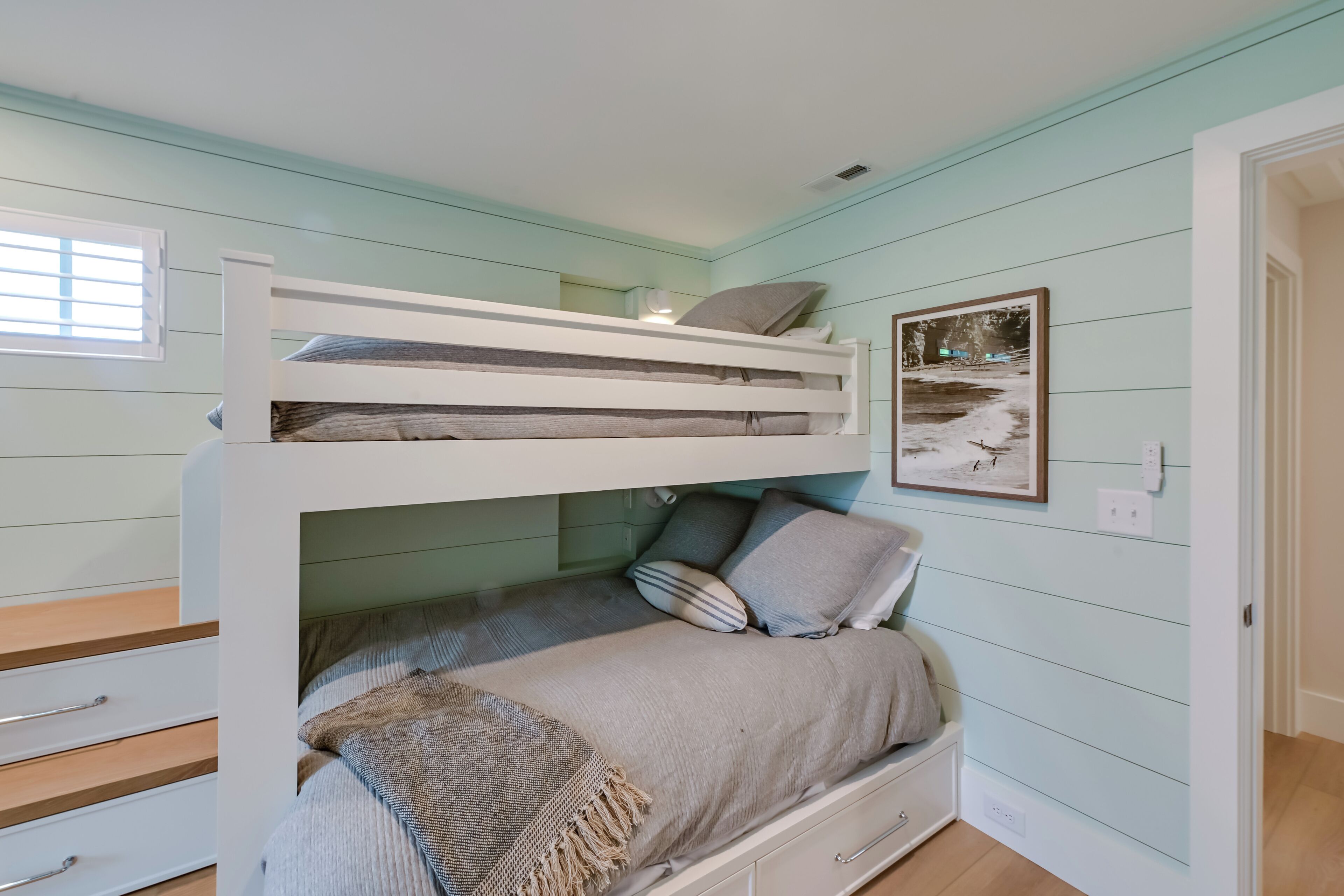 A bunk bed room with a window, a framed picture on the wall, and a door leading to another room.