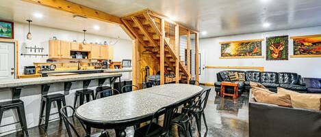 A spacious living area with a large round table, black chairs, a leather sofa, and a staircase leading to an upper level.