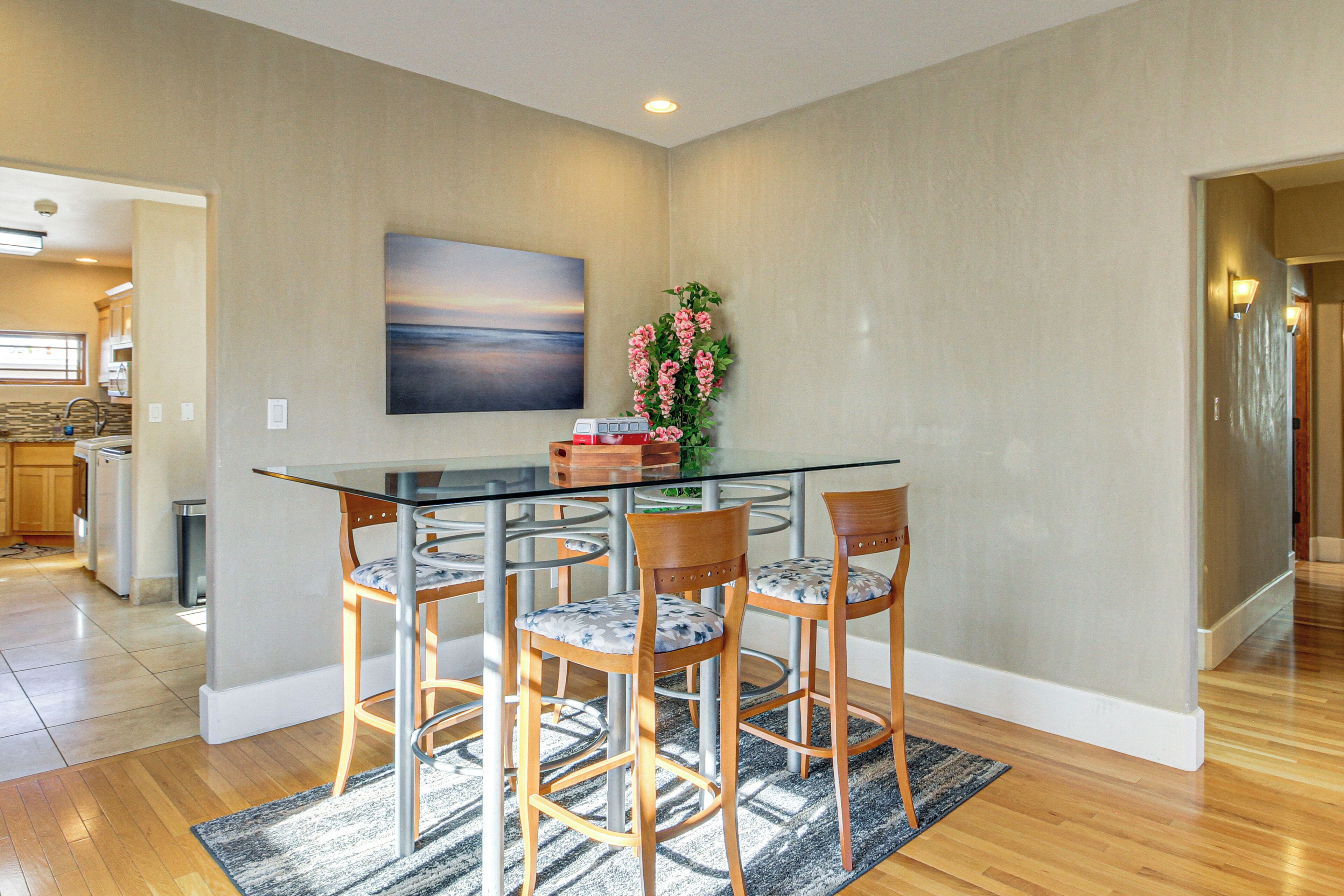 A dining area with a glass table, four wooden chairs, a vase with flowers, and a painting on the wall.