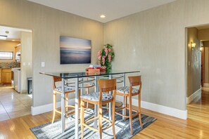 A dining area with a glass table, four wooden chairs, a vase with flowers, and a painting on the wall.