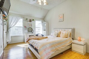 A bedroom with a bed, bedside tables, a dresser, a window with curtains, a ceiling fan, and a wall-mounted shelf with decorative items.