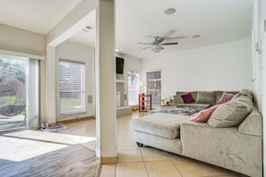 A spacious living room with a large sofa, a ceiling fan, and a view through a sliding glass door to the outdoors. - Arlington Home: Pool Oasis Near At&t Stadium (Grand Prairie)