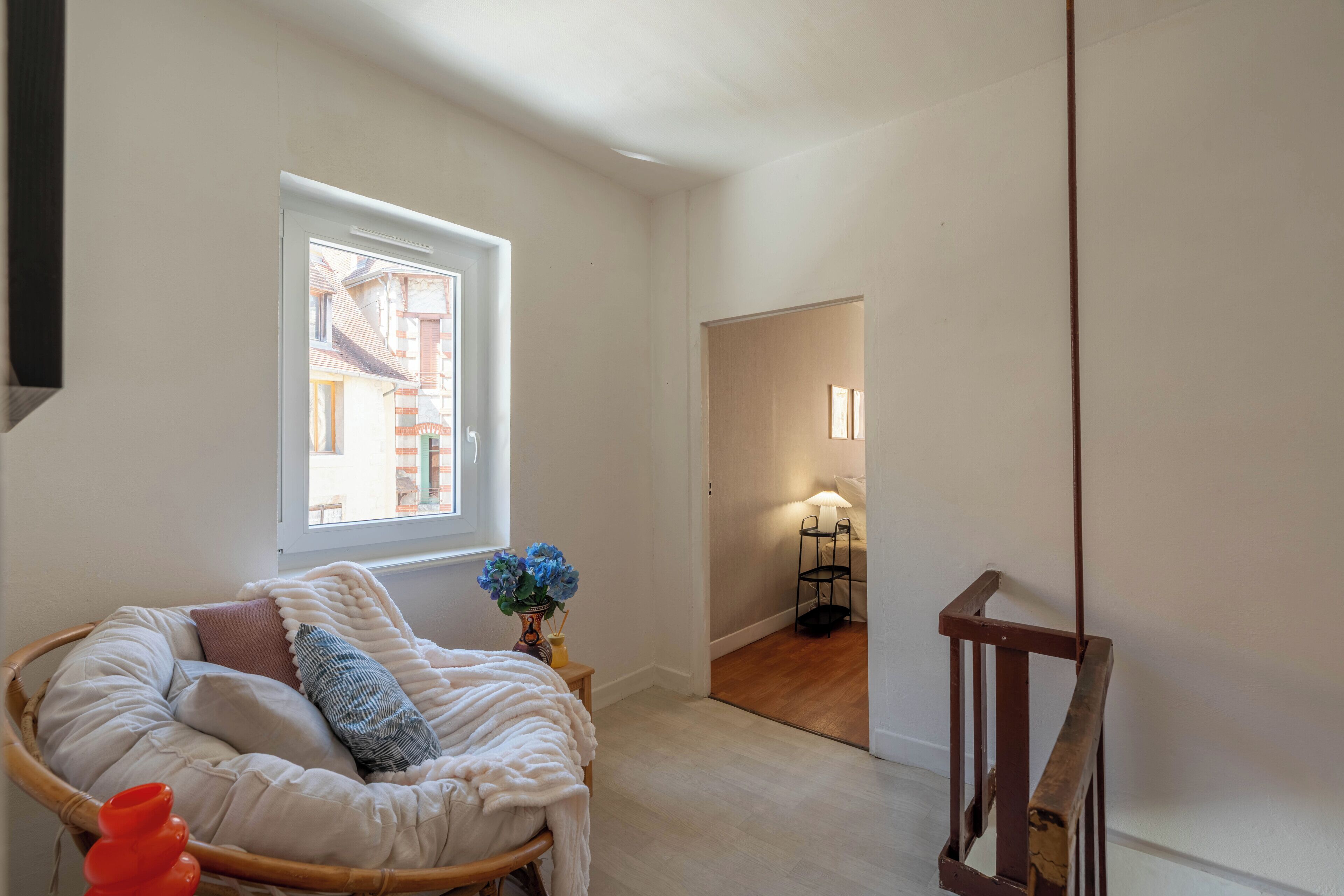 Quarto com cama de vime, janela com vista, mesa pequena com um vaso e uma luminária.