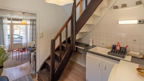 Una escalera conduce a un segundo piso, una cocina con armarios blancos, un comedor con mesa y sillas, y un ventanal con cortinas.