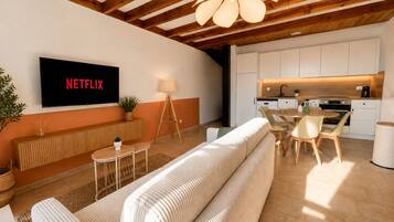 A modern living room with a sofa, a TV displaying 'NETFLIX,' a dining area with chairs, and a kitchen with white cabinets.