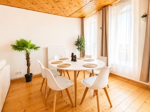 A round dining table with white chairs, a potted plant, and a radiator in a room with wooden flooring and a wooden ceiling.