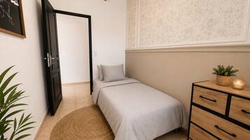 Un dormitorio con cama, cómoda, alfombra redonda tejida, planta en maceta y pared con estampado floral.