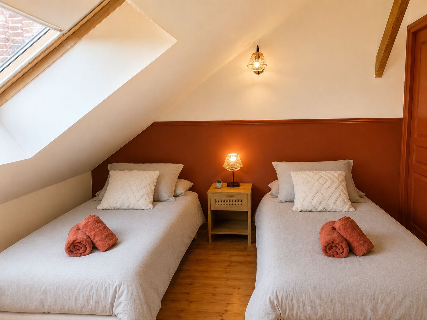 A bedroom with two beds, a skylight, a nightstand with a lamp, and a wooden floor.