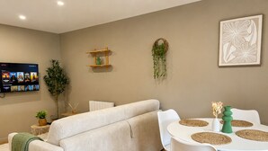 A living room with a sofa, a dining table with chairs, a TV mounted on the wall, and a plant on the floor.