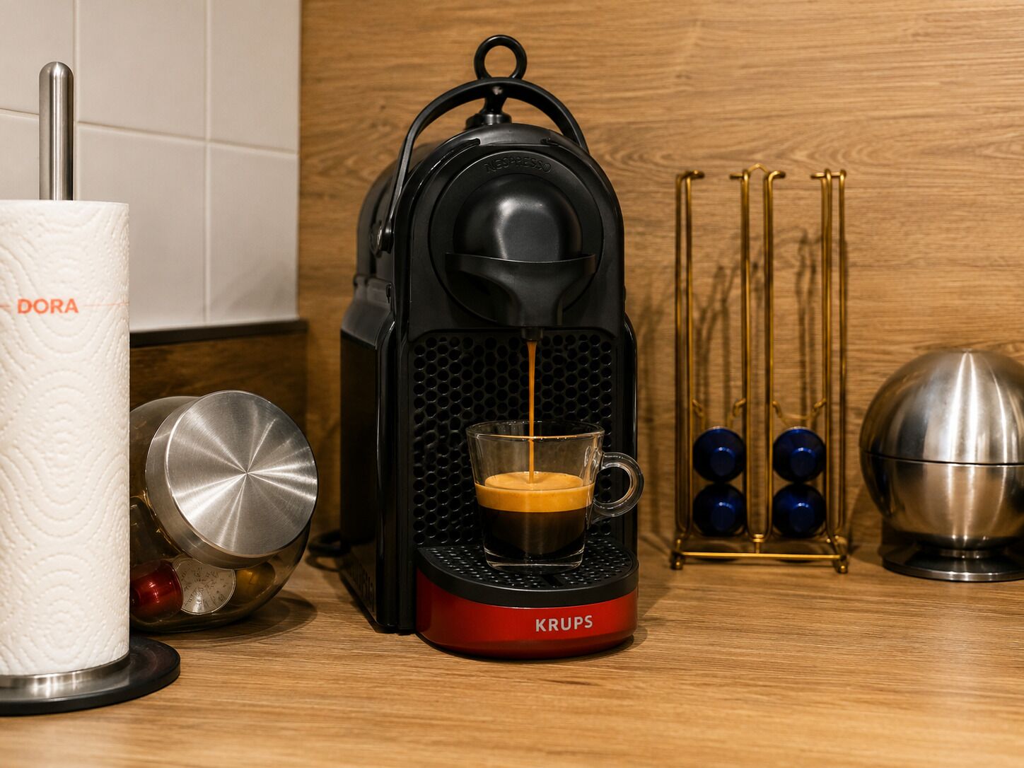 Una cafetera Krups con una taza de vidrio en su base, un rollo de pañuelos y objetos decorativos sobre una superficie de madera.