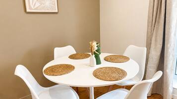 Une table ronde blanche, entourée de quatre chaises blanches, chacune agrémentée d’un set de table en fibres tressées. Un petit vase blanc contenant des fleurs est posé sur la table.