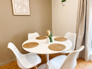 A round white table with four white chairs, each with a woven placemat. A small white vase with flowers is on the table. - Apart'Moment - Résidence La Brasserie (Aire-sur-la-Lys)