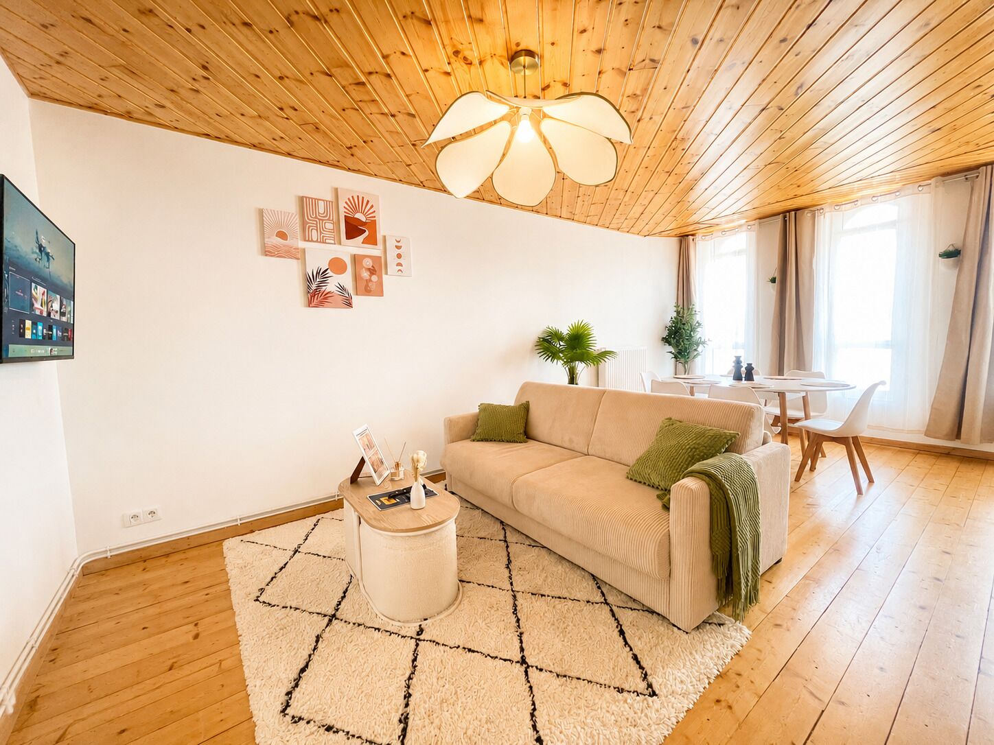 Een woonkamer met een houten plafond, een beige bank, een kleine ronde tafel en een eethoek met een witte tafel en stoelen.