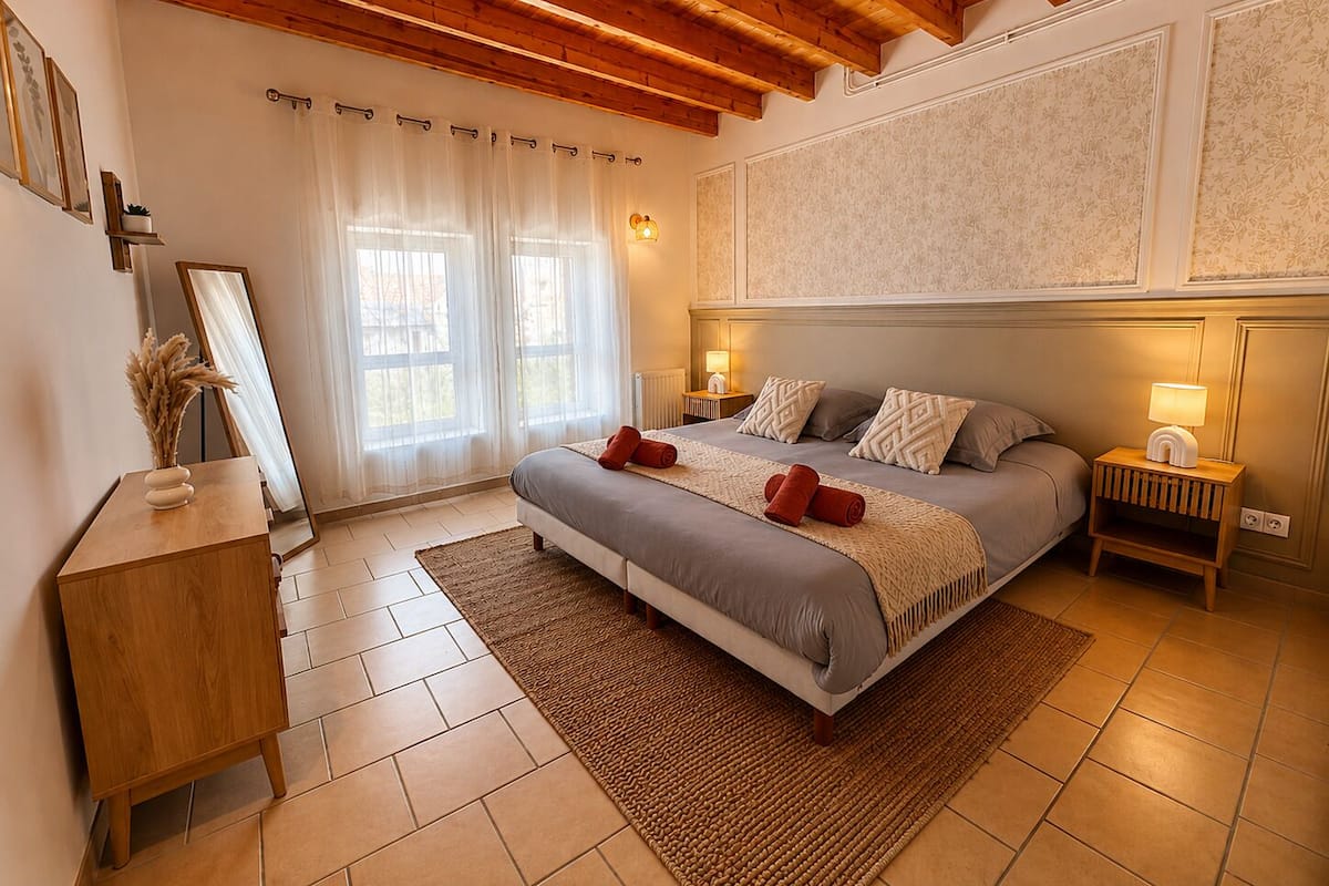 A bedroom with a bed, bedside tables, a mirror, a wooden dresser, and a window with sheer curtains.