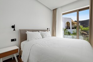 A bedroom with a large bed, a bedside table, a wall-mounted lamp, and a view of a building and greenery through a window.