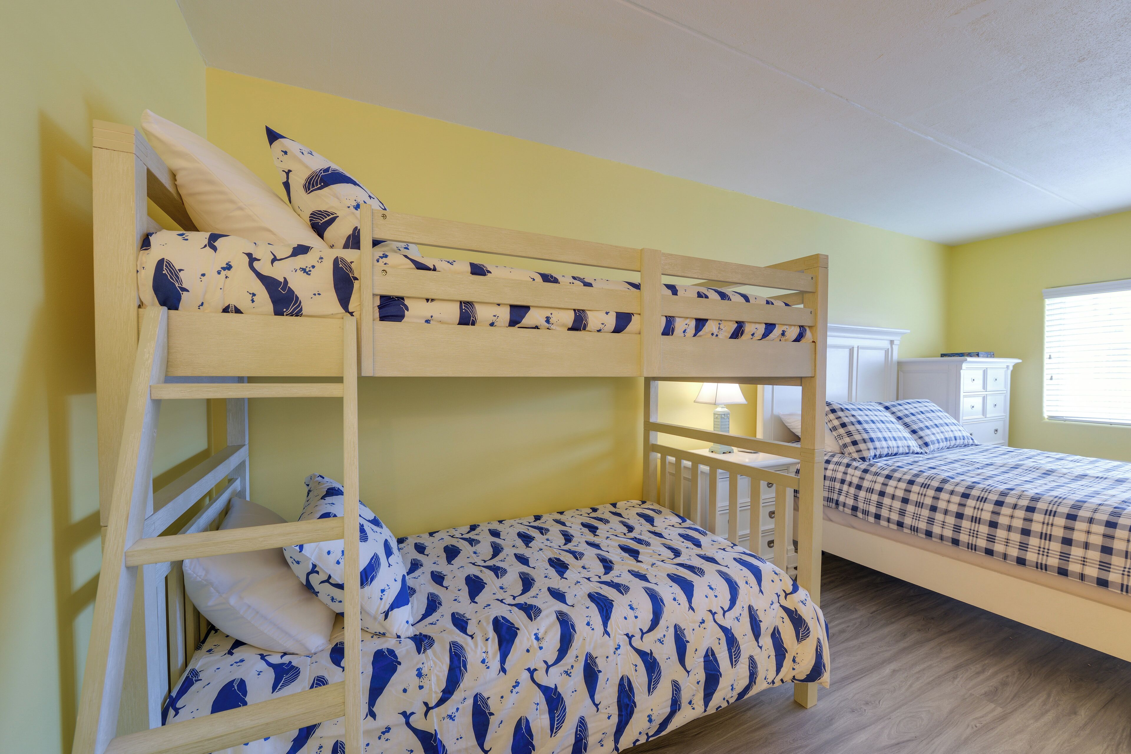A bunk bed room with a yellow wall, a wooden floor, and a window with blinds.
