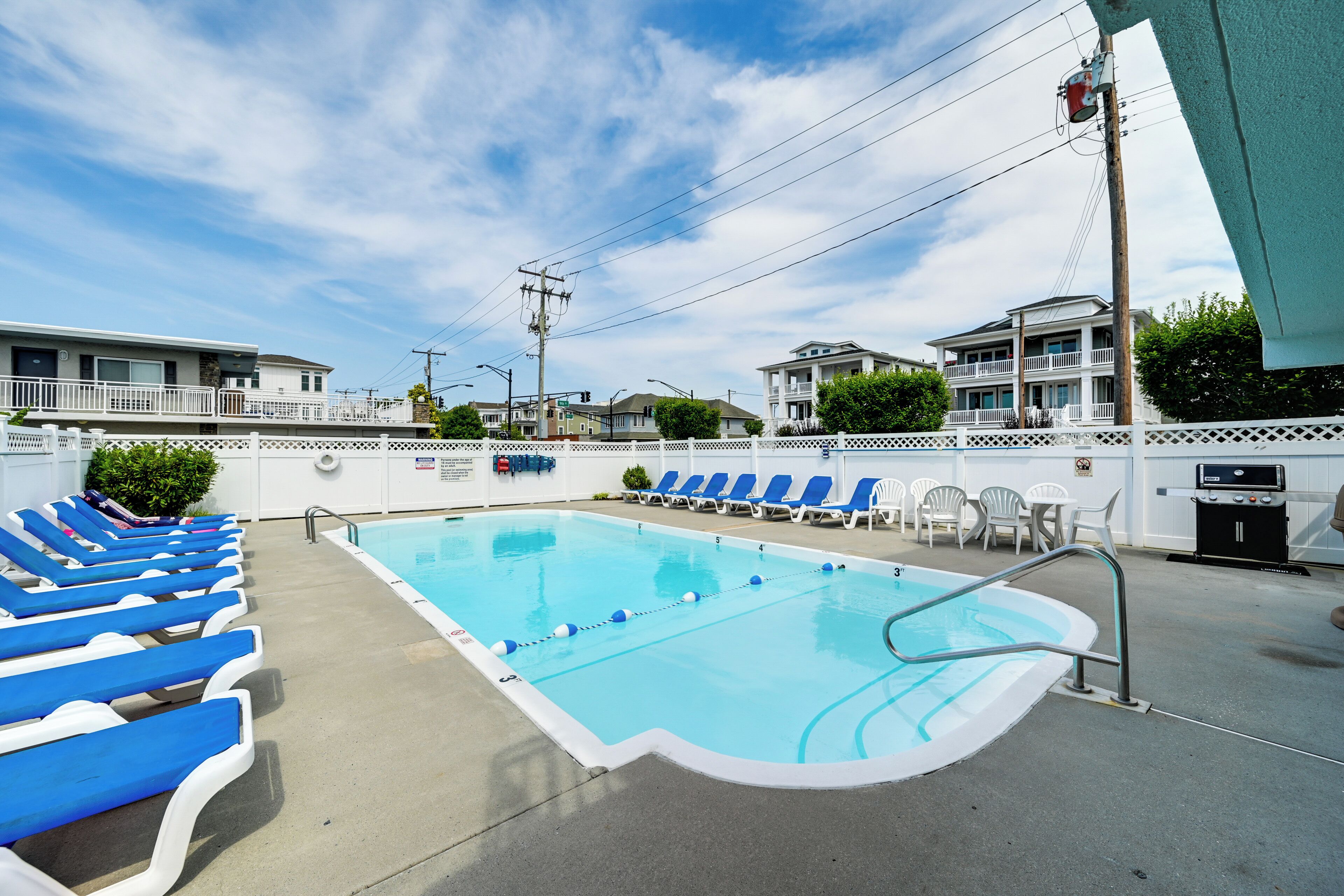 A swimming pool with blue lounge chairs, a white fence, and a building in the background.