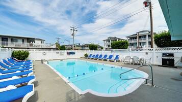 A swimming pool with blue lounge chairs, a white fence, and a building in the background.