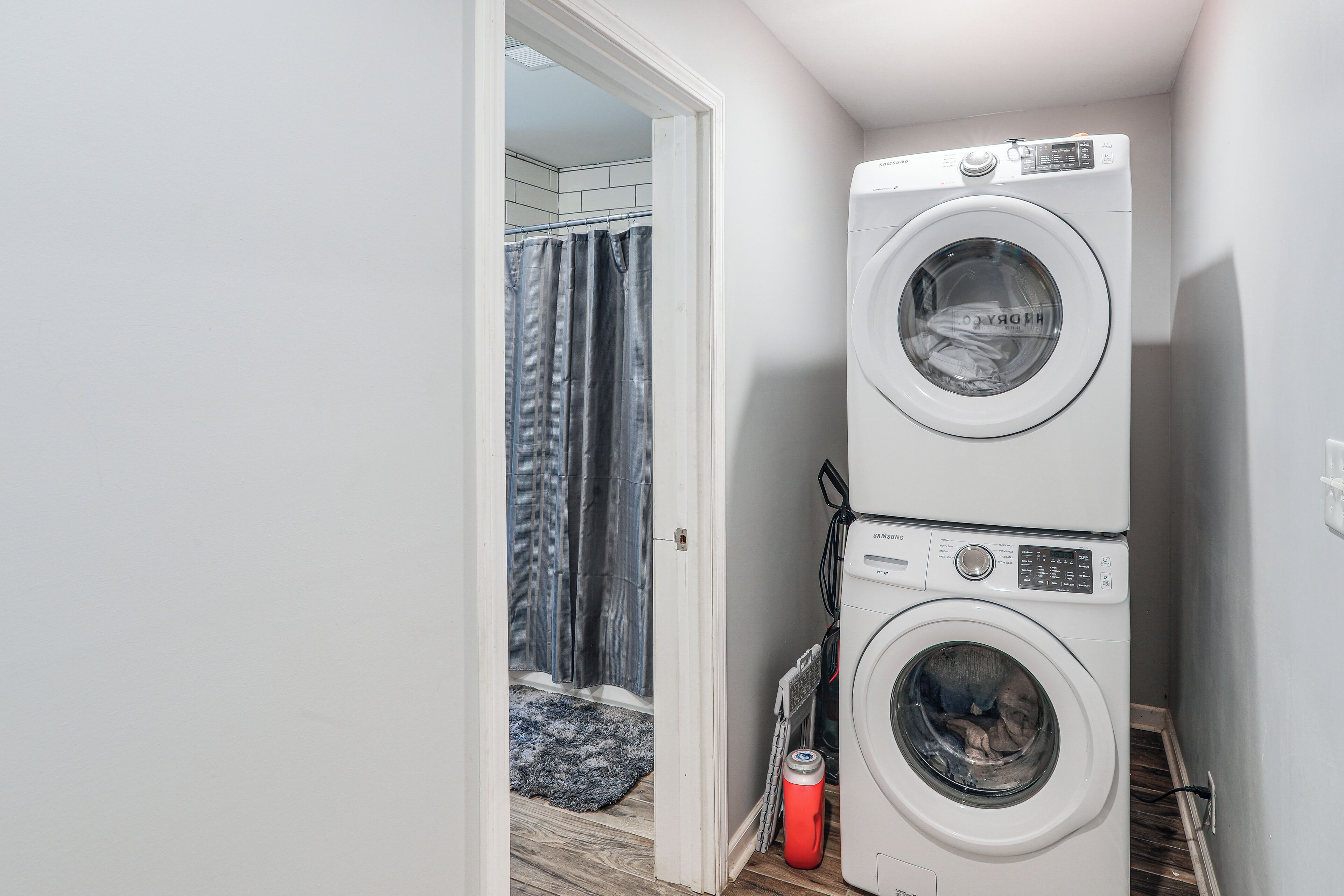Ein Waschraum mit zwei übereinander gestapelten Waschmaschinen, einer Duschvorhang und einem roten Warmwasserbereiter.