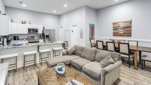 A modern kitchen with white cabinets, a dining area with a wooden table and chairs, and a living room with a beige sectional sofa.