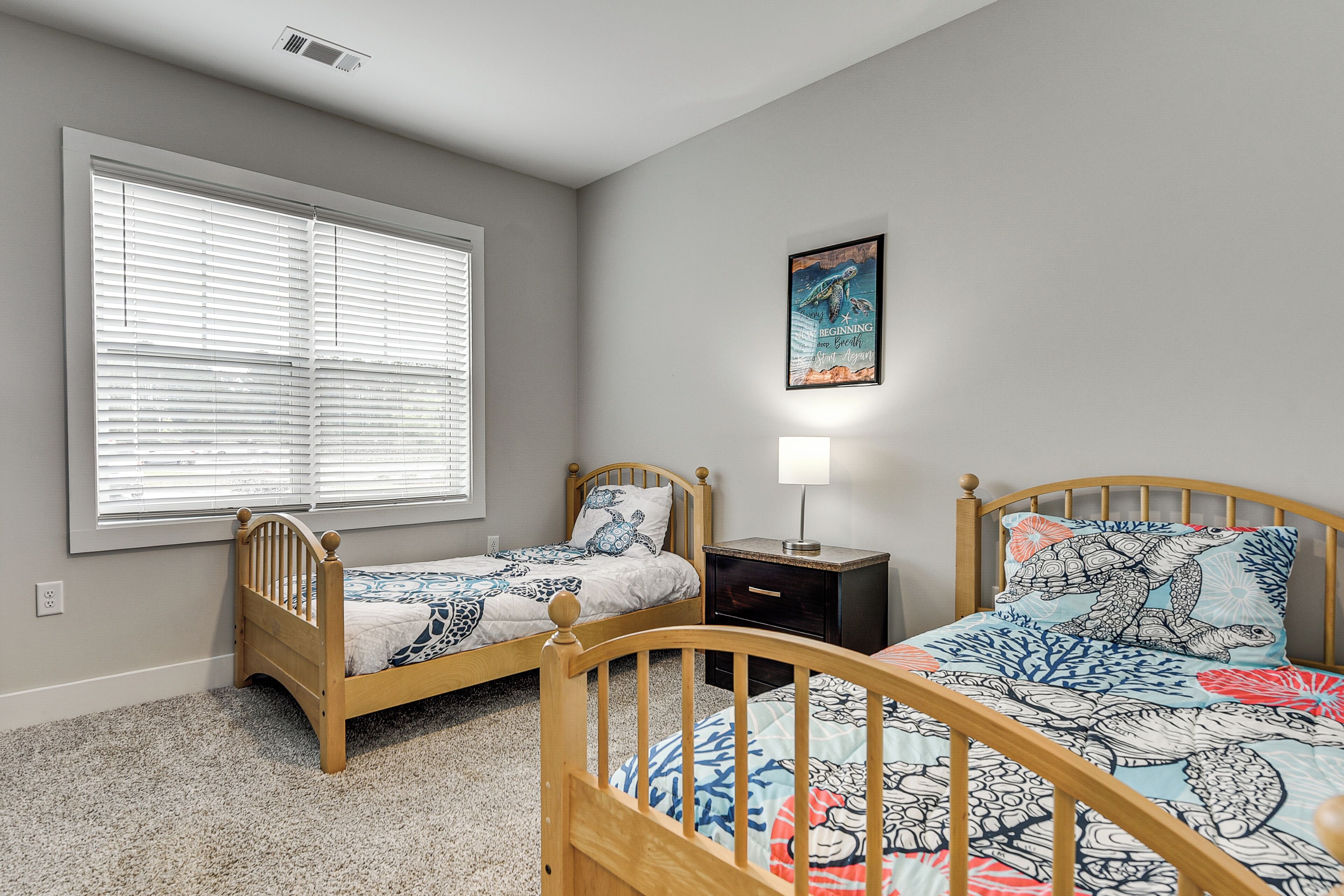 A bedroom with two wooden beds, a nightstand, a wall-mounted lamp, and a large window with blinds.