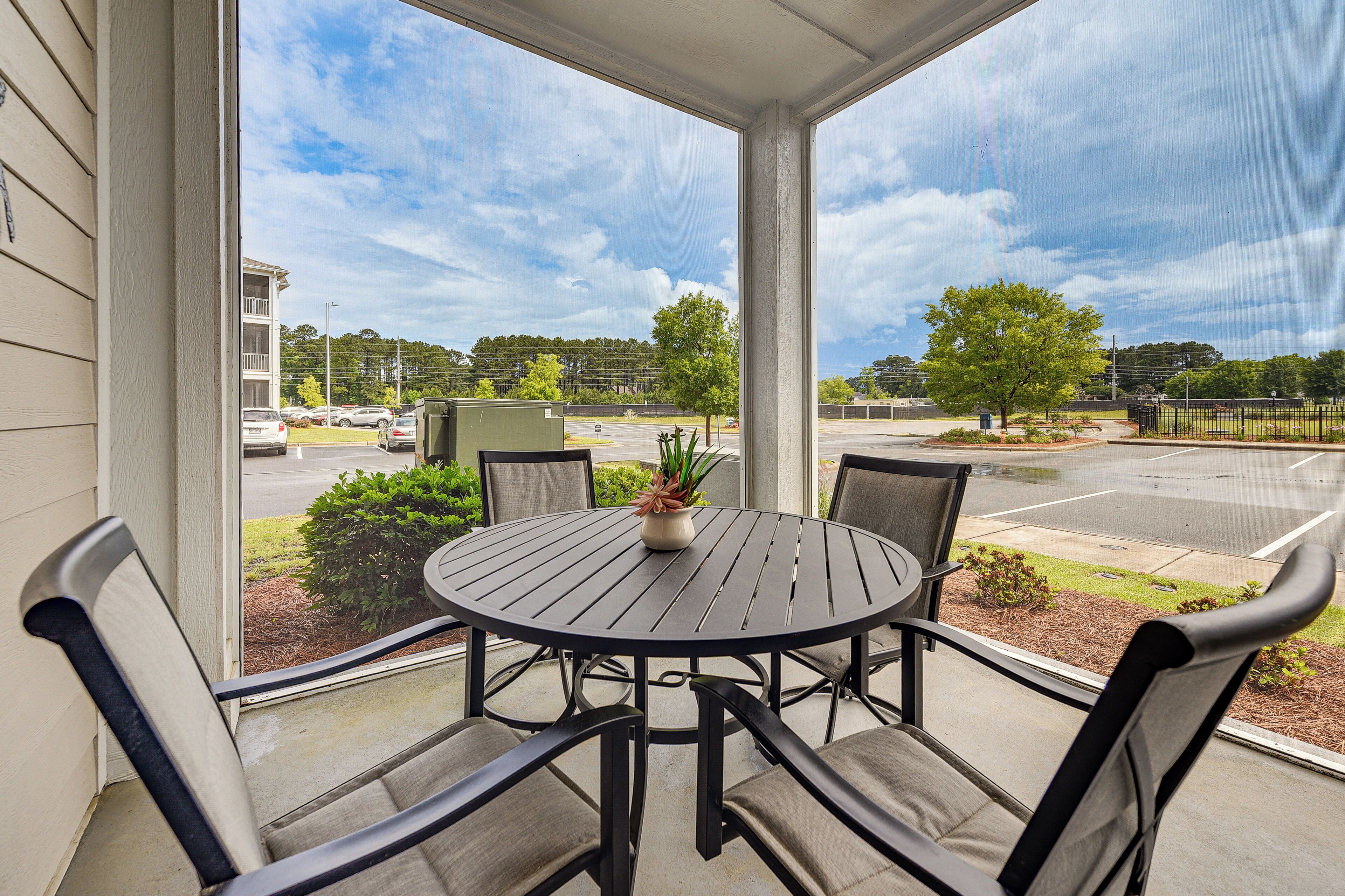 Une terrasse couverte avec une table ronde et quatre chaises, un pot de fleurs et une vue sur un parking et des arbres.