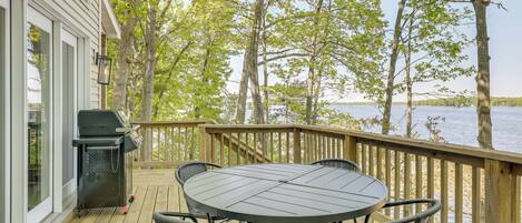 A wooden deck with a round table and chairs, a grill, and a view of a lake.
