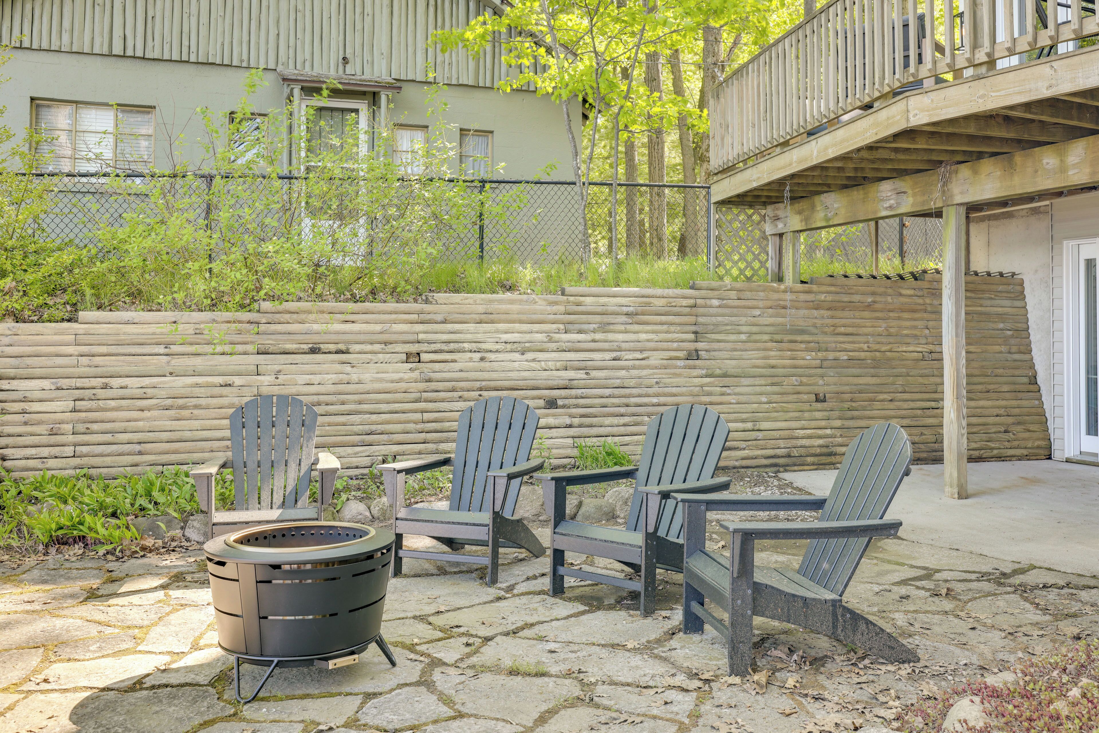 A patio with Adirondack chairs and a fire pit, a wooden fence, and a building in the background.