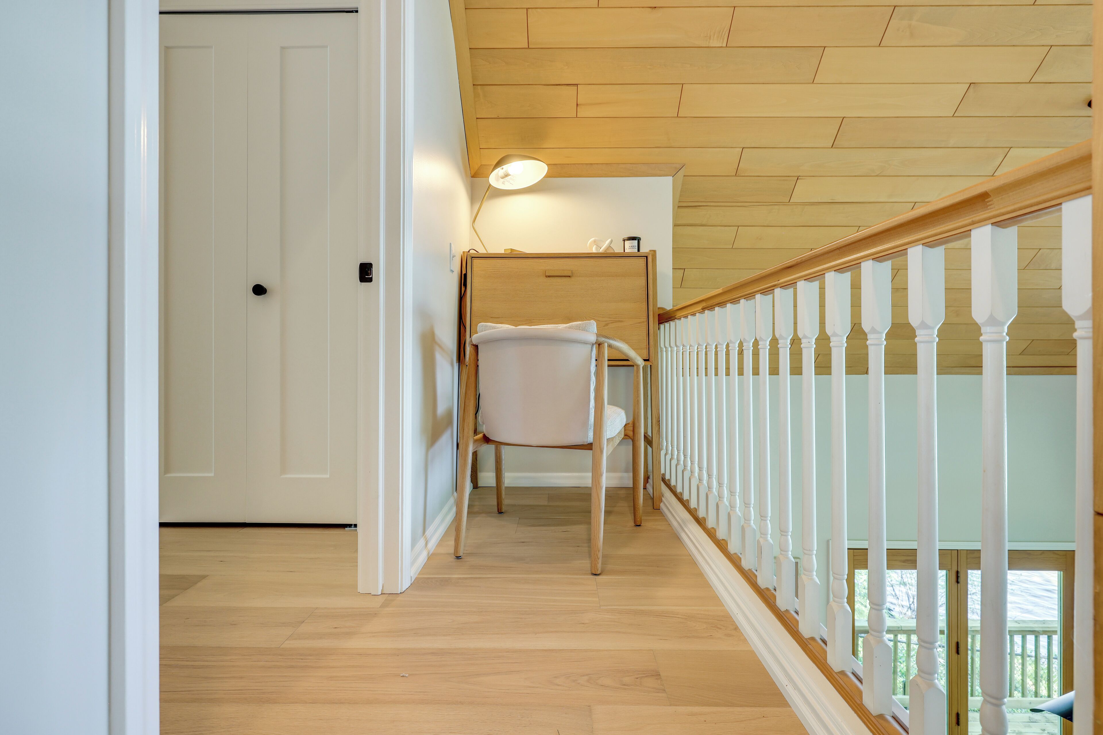 A wooden-floored hallway with a white chair, a white door, and a railing with balusters.