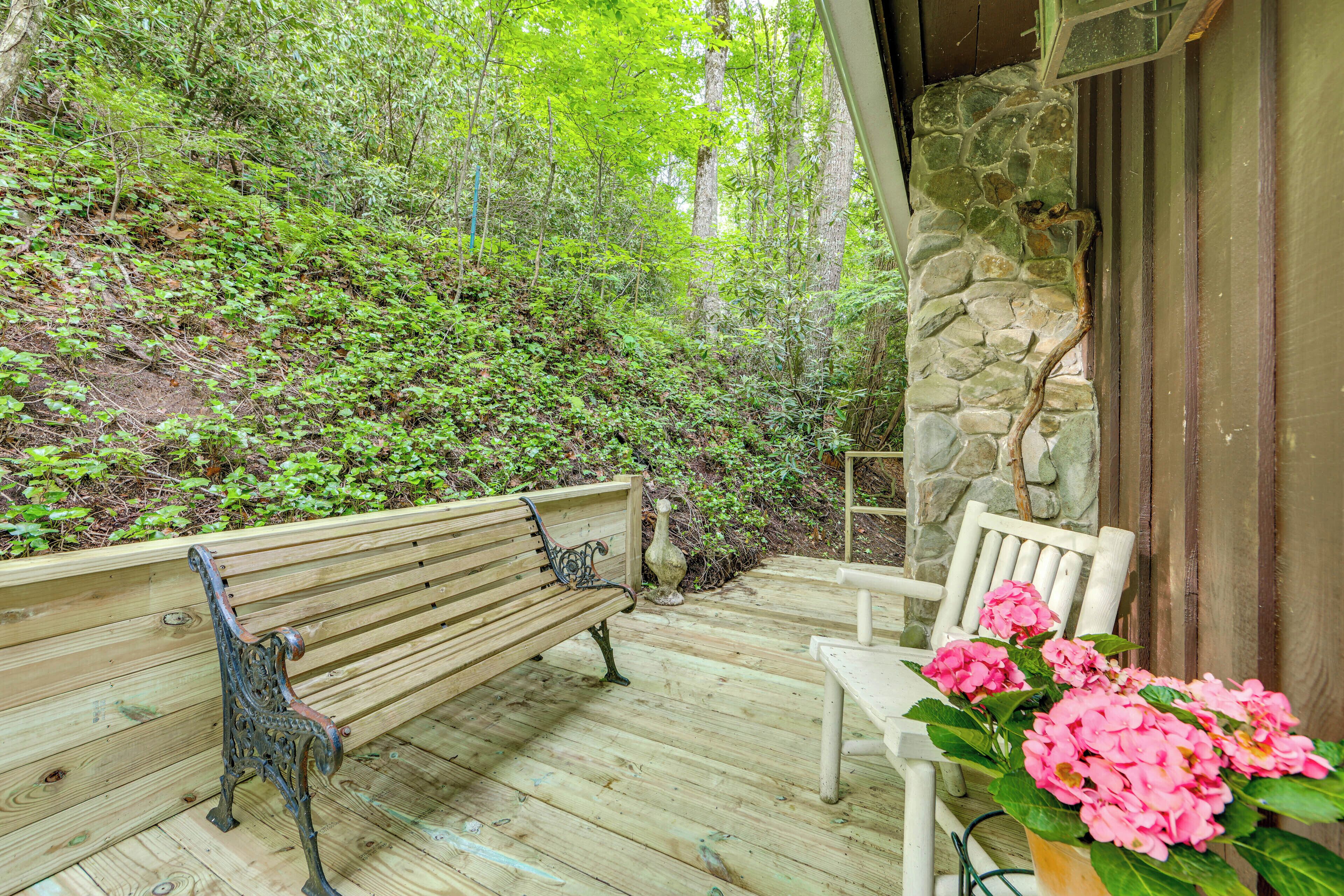 Une terrasse en bois avec un banc et une chaise, entourée de verdure et d’un mur de pierres.