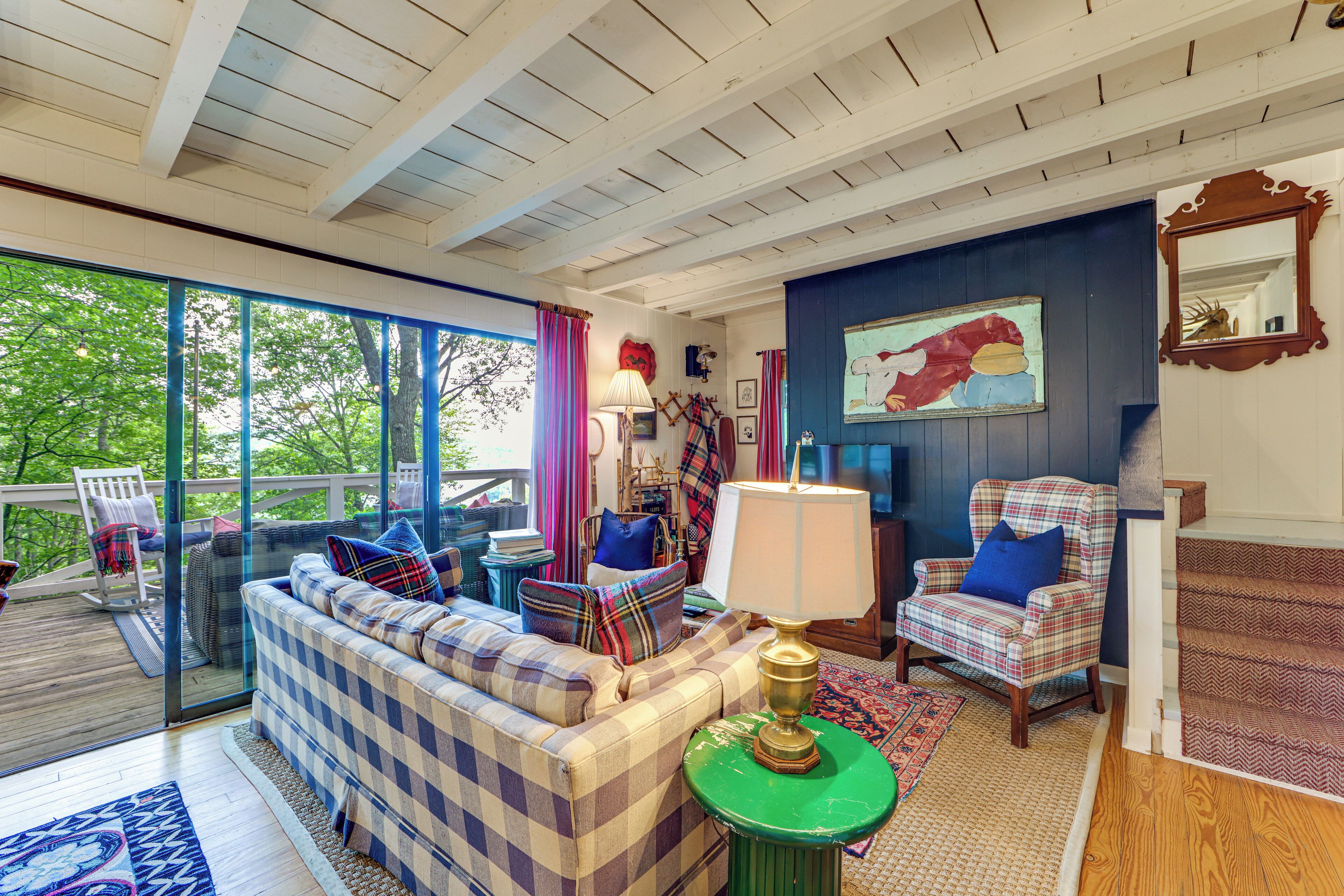 A cozy living room with a checkered sofa, a plaid armchair, a green side table, and a large window overlooking greenery.