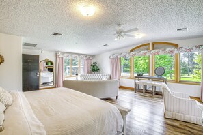 A bedroom with a large bed, a sitting area with a chair, a vanity, and a view of greenery outside.