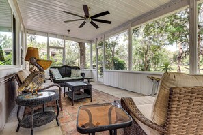 A covered porch with wicker furniture, a ceiling fan, and large windows overlooking a wooded area.