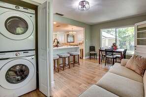 A laundry room with a front-loading washer and dryer, a dining area with a chandelier, and a living room with a sofa and a window.