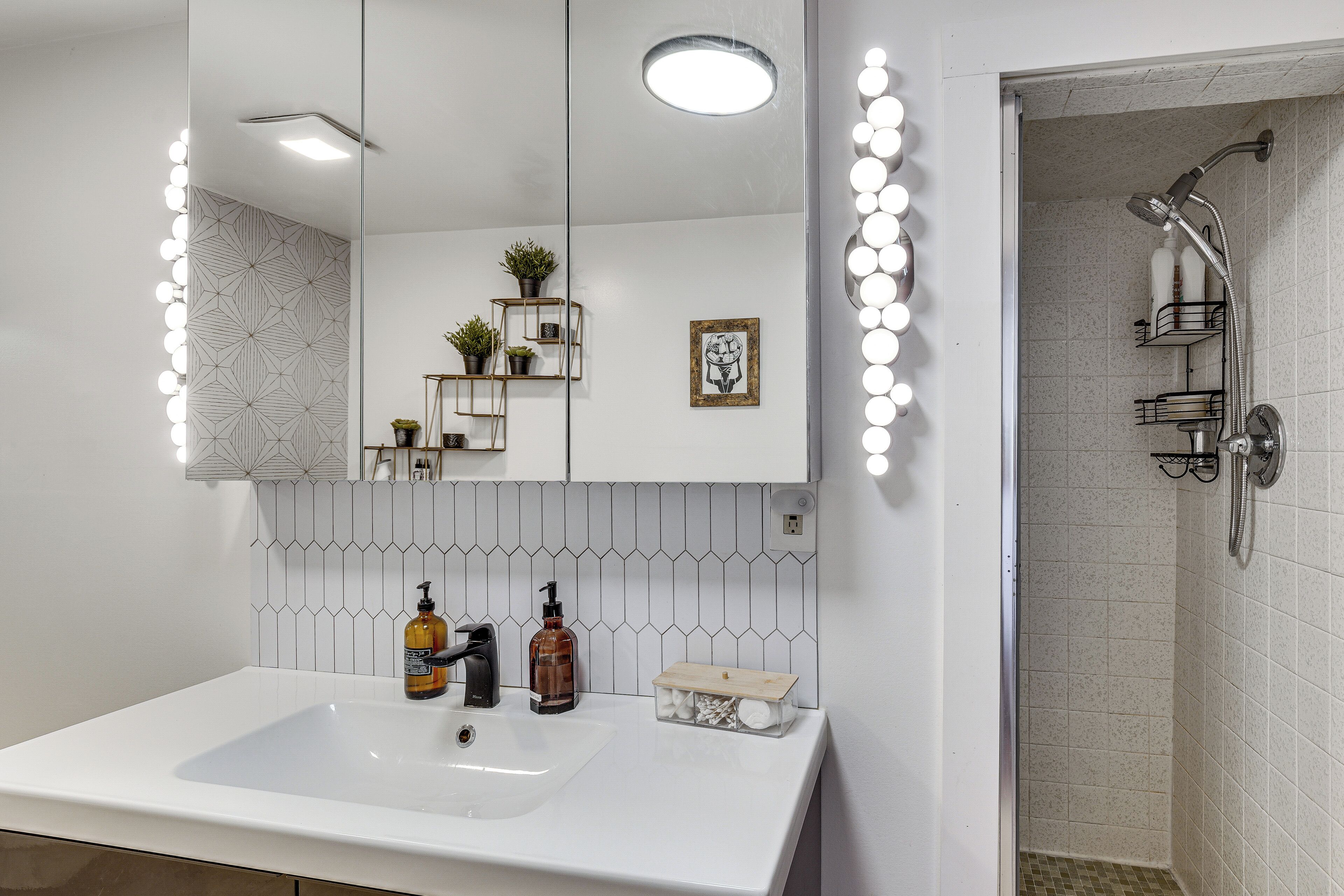 A bathroom with a white sink, a mirror with lights, a shower, and a tiled wall.