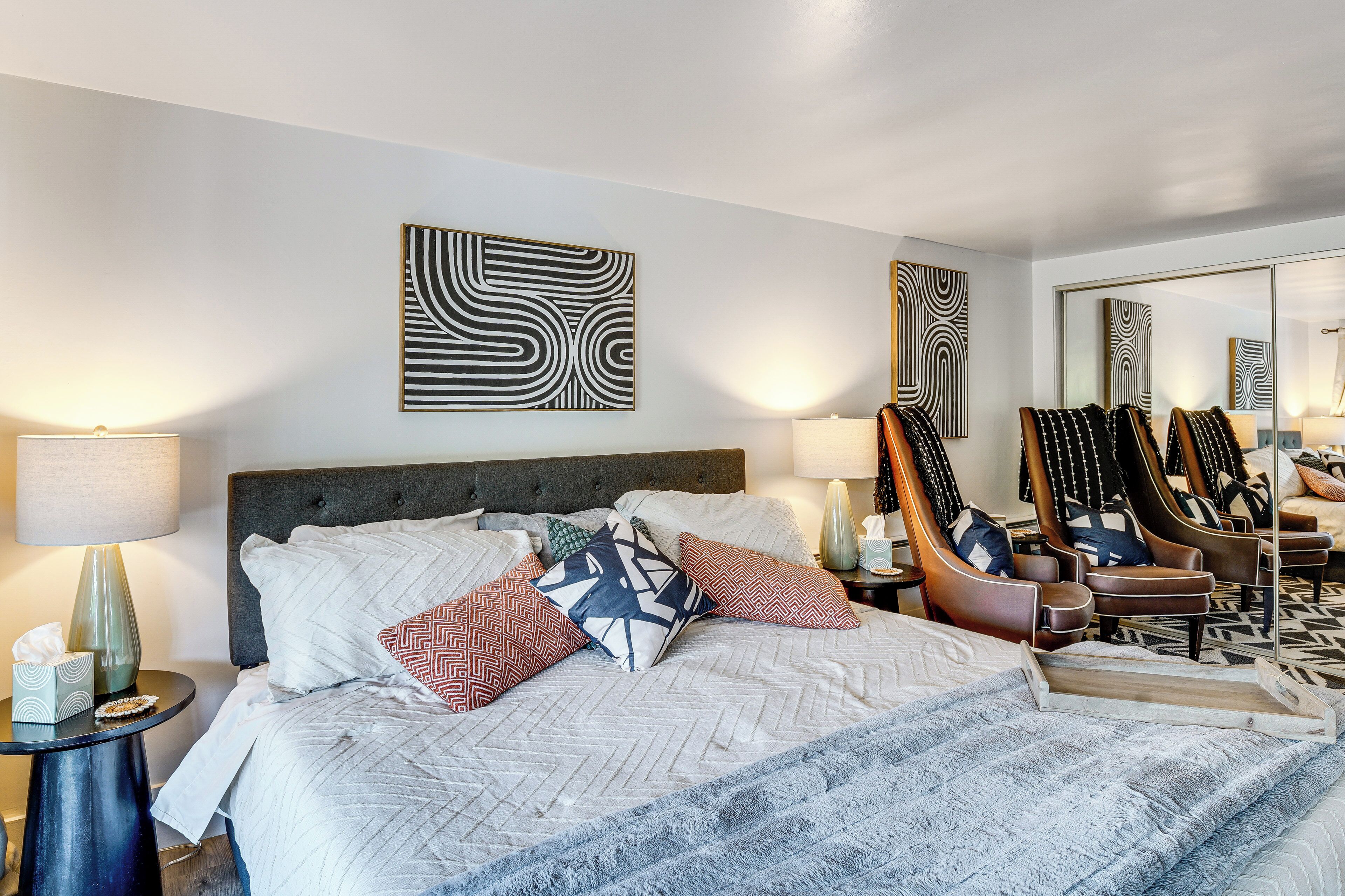 A hotel room with a large bed, multiple pillows, a headboard, a side table with a lamp, a mirror, and a seating area with chairs.