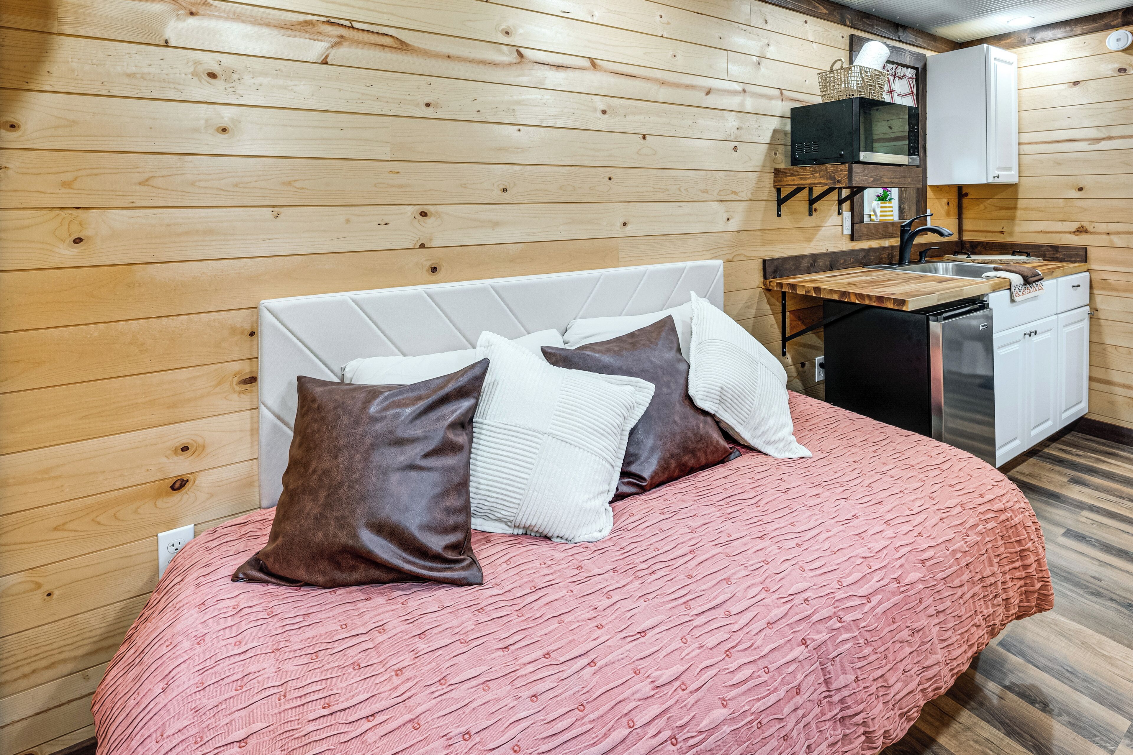 Un dormitorio con cama, almohadas, pared de madera, una pequeña kitchenette con lavamanos y microondas.
