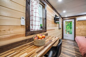 A wooden interior with a wooden bench, a basket of fruit, a chair, and a bed with a pink blanket.