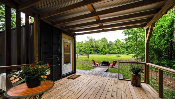A wooden deck with a table and chairs, a door leading to a covered area, and a view of a grassy yard.
