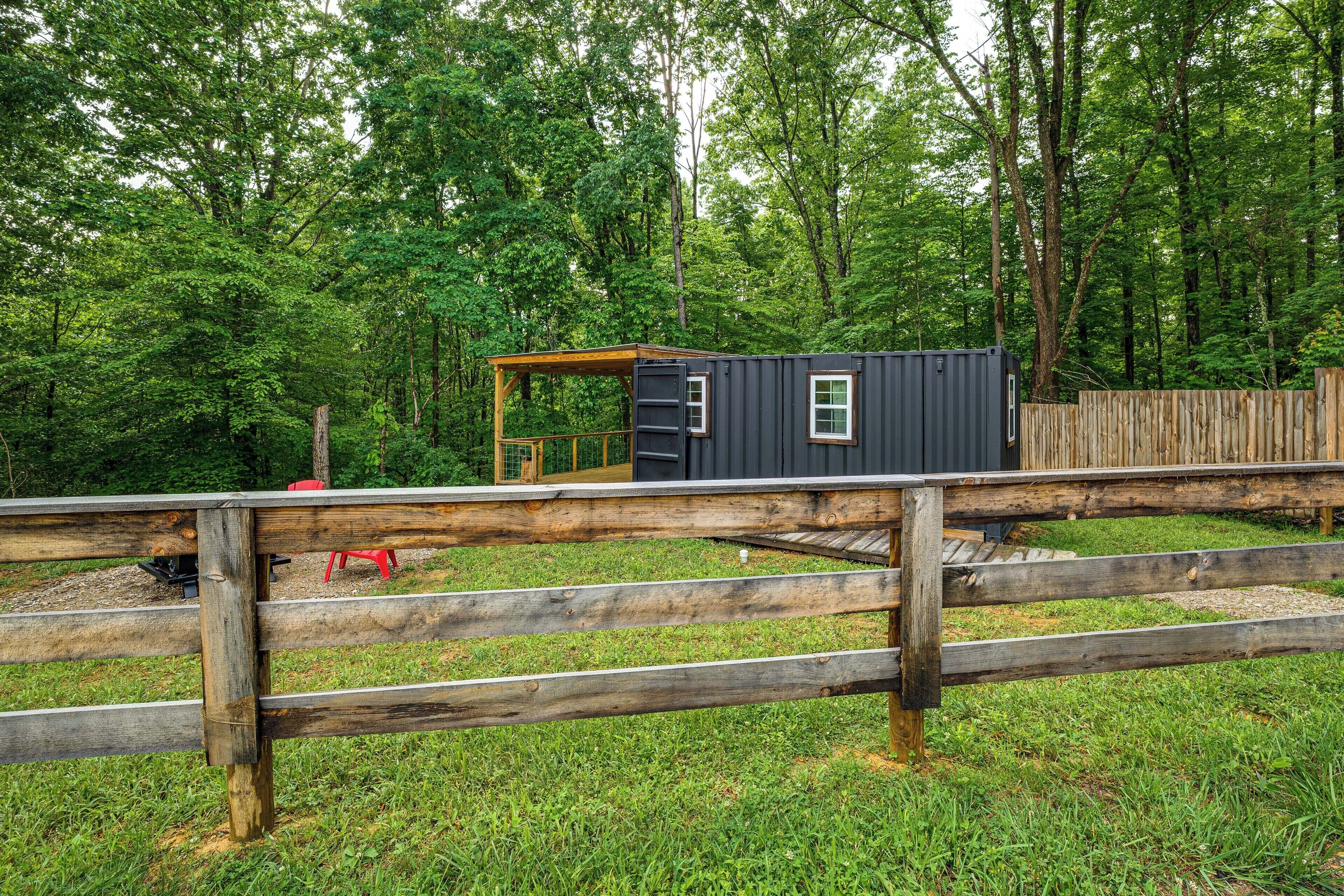 A small, dark-colored cabin with a wooden deck and a fenced area in a forested setting.