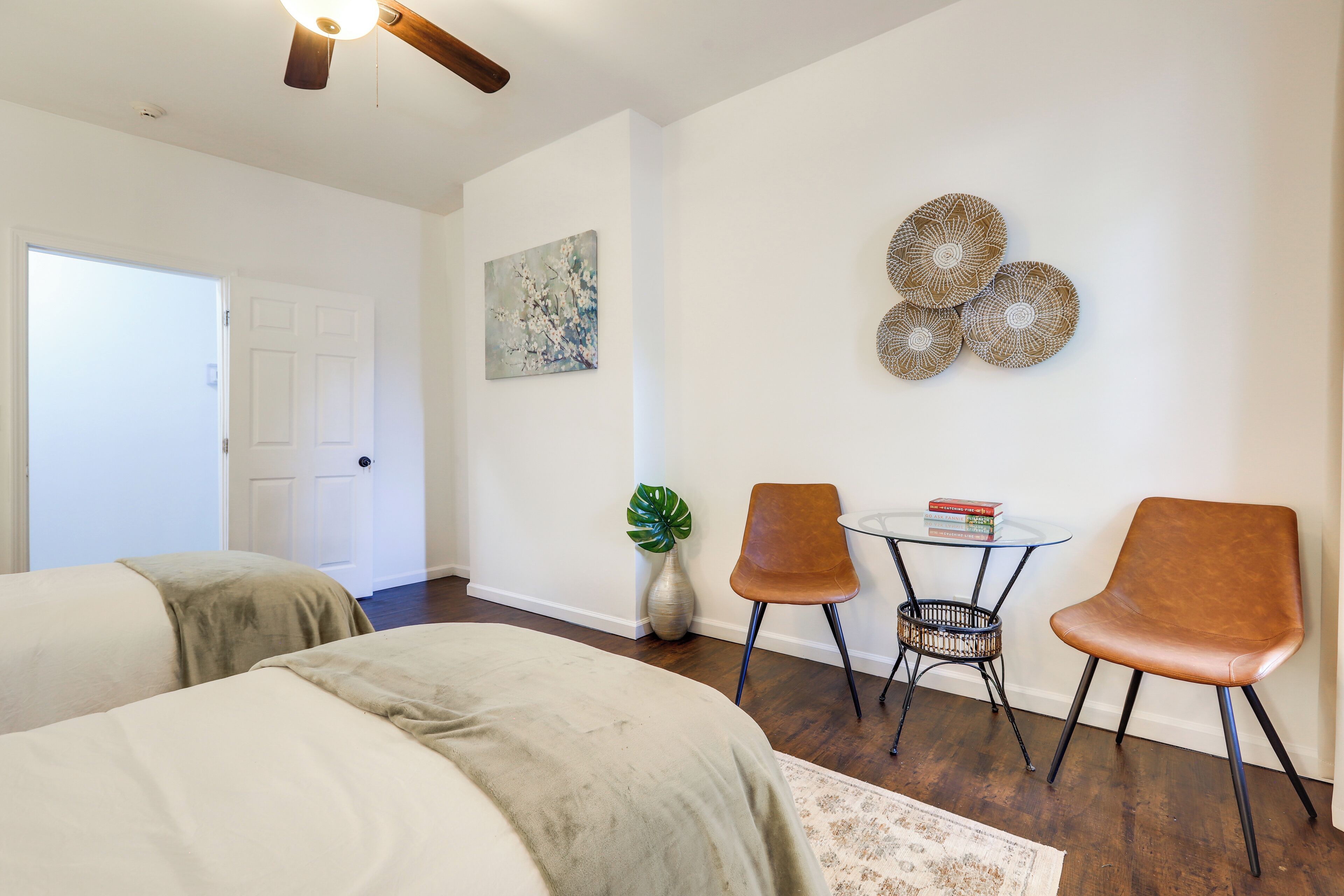 A bedroom with two beds, a ceiling fan, a door, a wall-mounted artwork, a plant, and a small table with chairs.