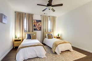 Ein Schlafzimmer mit zwei Betten, einem Deckenventilator, beigen Vorhängen, einem Gemälde, Nachttischlampen und einem Teppich.