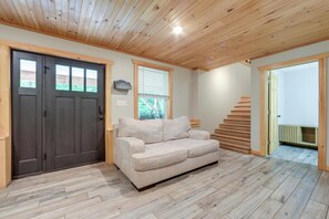 A room with a wooden ceiling, a dark door, a beige sofa, a window with blinds, and a staircase leading to another room.