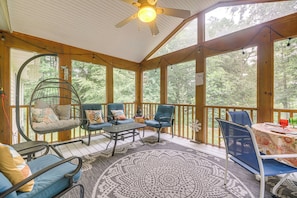 A covered porch with wooden beams, a ceiling fan, and a large window overlooking a forest.