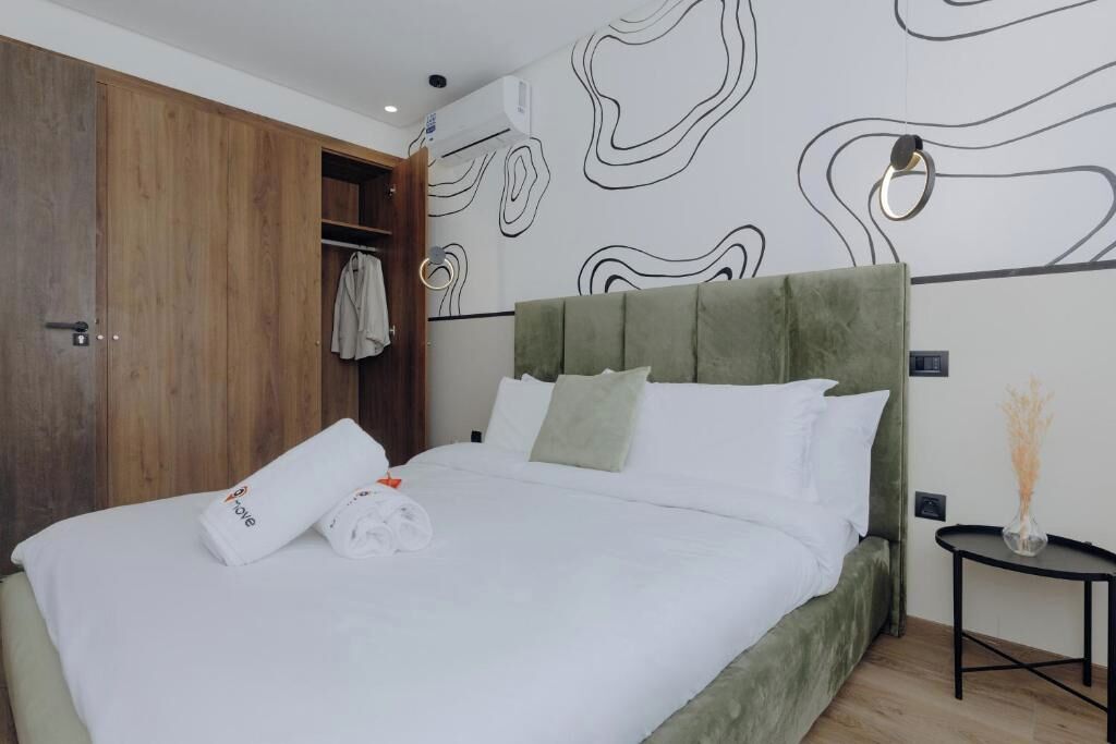 Une chambre à coucher avec un lit recouvert d’un tissu vert, du linge de lit blanc, une armoire en bois et des œuvres d’art abstraites accrochées au mur.