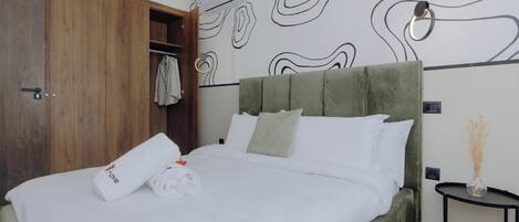 Une chambre à coucher avec un lit recouvert d’un tissu vert, du linge de lit blanc, une armoire en bois et des œuvres d’art abstraites accrochées au mur.