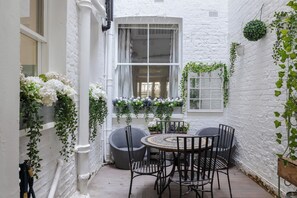 A white brick courtyard with a round table and chairs, a window with curtains, and hanging plants. - Stunning Notting Hill Flat (London)