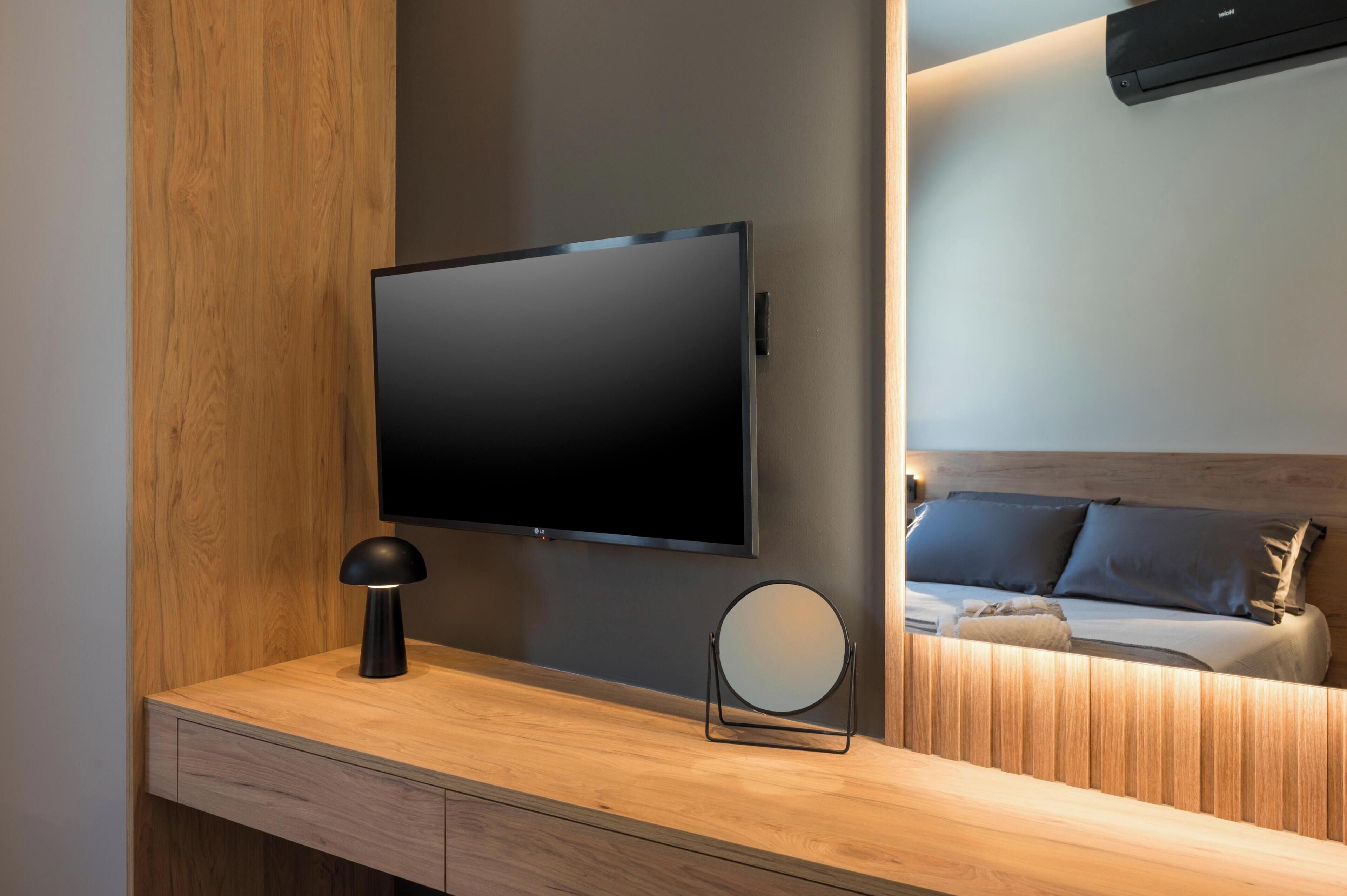 A modern room with a flat-screen TV mounted on the wall, a bedside table with a lamp, a round mirror, and a bed with a headboard.