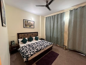 Une chambre à coucher avec un lit, un ventilateur de plafond, une table de chevet, un tableau encadré au mur et une fenêtre avec des rideaux.