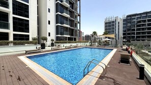 A rectangular swimming pool with a metal ladder on one side, surrounded by a wooden deck. - Rentico l Luxury 1BR Pool View l Azizi Riviera (Dubai)